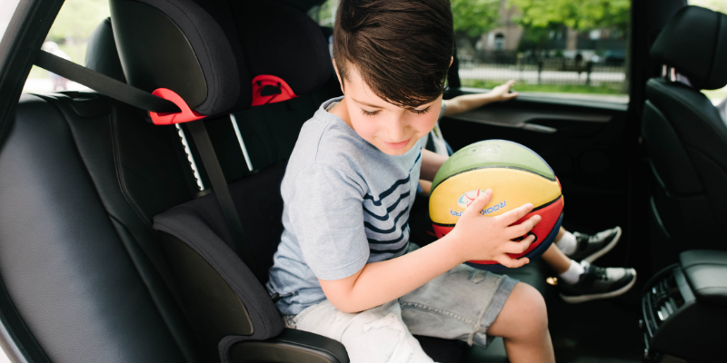 Little boy getting out of his Clek Oobr Booster Seat holding a basket ball.