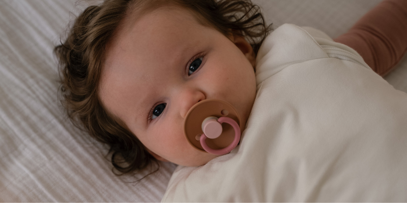 Baby lying in Crib with Pip + Phee Sleep Sack in Sand Color on and Soother in Mouth
