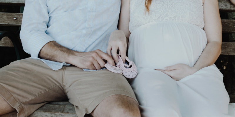 Man and Woman Sitting Down As Woman Holds Her Pregnant Stomach and Both Are Holding a Pair of Baby Shoes