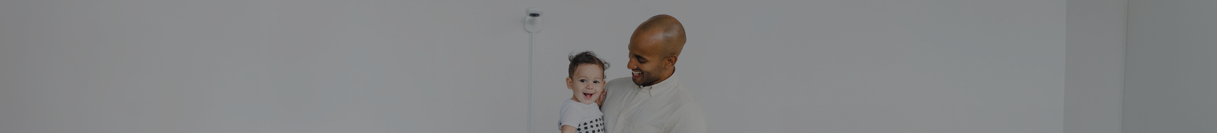 Father holding baby in his nursery wearing a Nanit breathing band while Nanit baby monitor is affixed to wall behind them