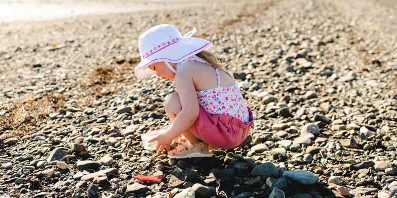 Toddler sitting on a rocky beach playing and wearing a sun hat