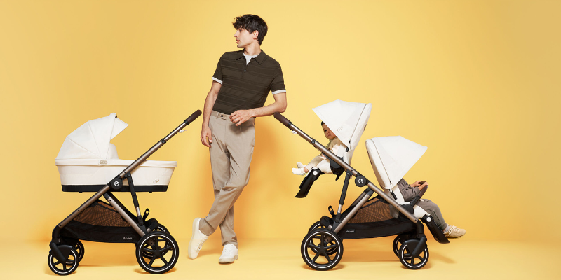 Man standing beside two Cybex Gazelle S 2023 Strollers, one with a bassinet attached and one with a toddler and second seat attached