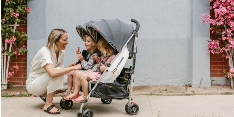 Mom kneeing down to talk to two little girls sitting in UPPAbaby G-link v2 double umbrella stroller