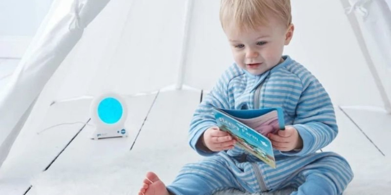 toddler in a white tent, dressed in blue, holding a book and next to a Gro Clock