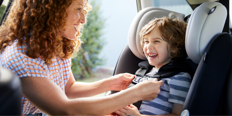 Maxi-Cosi Pria Convertible Car Seat With Little Boy Sitting Inside Smiling While Woman Buckles Him In