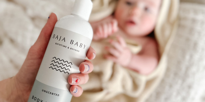 Baby Wrapped Up in Towel Lying Down  With Adult Holding Baja Baby Lotion Infront of Them