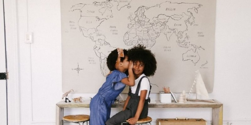 Little Girl Whispering in Other Little Girls Ear With Picture of Map in Background