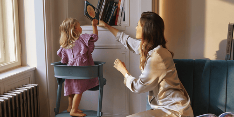 Little Girl Standing in Cybex LEMO Learning Tower Set Grabbing A Book While Woman Helps Her