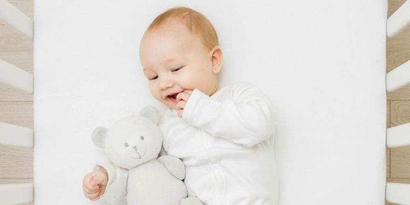 Baby laying in a crib on a Simmons Baby Breathable Crib Mattress holding a grey bear