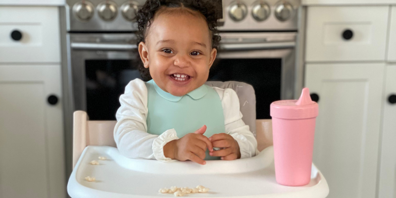 Child Sitting in High Chair Smiling With Pink Sippy Cup Beside Her