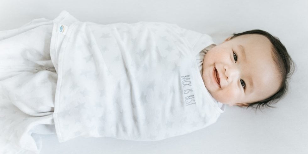 Smiling baby in HALO Sleep Swaddle embroidered with "Back is Best"