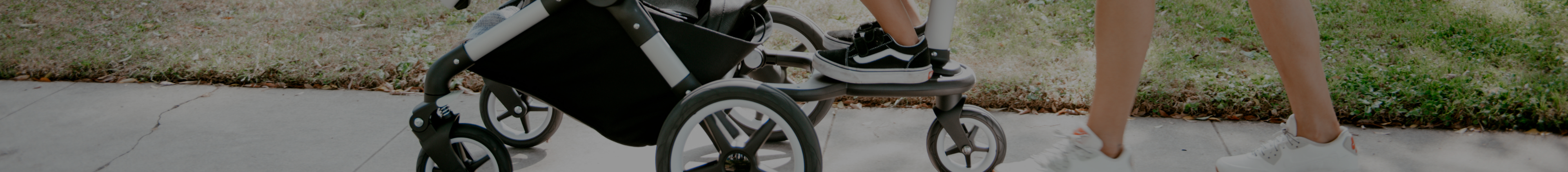 Child standing on a stroller board while mother pushes Bugaboo stroller in the park.